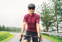 Дорослий спортсмен у велосипедних сонцезахисних окулярах і шоломі сидить на дорожньому велосипеді на сільській дорозі вдень — стокове фото