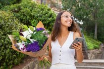 Content young female in eyeglasses with blossoming flower bouquet text messaging on cellphone on urban stairs — Stock Photo