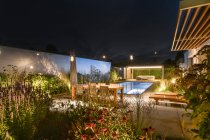 Просторный задний двор современной виллы с деревянным столом и шезлонгами и зелеными растениями, расположенными рядом с бассейном на фоне безоблачного ночного неба — стоковое фото