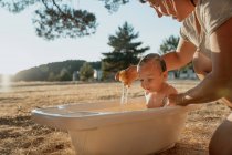 Ritagliato irriconoscibile mamma bagnando bambino mentre seduto sul prato sotto il cielo lucido nella giornata di sole — Foto stock