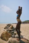 Vue latérale du contenu jeune femme afro-américaine en maillot de bain debout sur le rocher tout en regardant la caméra — Photo de stock