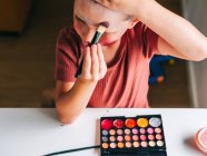 Criança encantadora com aplicador de maquiagem tocando a cabeça enquanto olha para a mesa com paleta de sombra — Fotografia de Stock