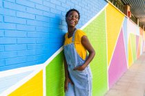 Бічний вид на щасливу дівчину афроамериканського походження, яка посміхається, стоячи на барвистій яскравій стіні. — стокове фото