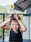 Fröhliches Kind in Gartenschürze mit blühendem Helianthus im Topf auf dem Kopf blickt auf Balkon in die Kamera — Stockfoto