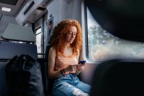 Заинтересованная женщина с вьющимися волосами в разорванных джинсах, отправляющая смс по мобильному телефону во время поездки на поезде днем — стоковое фото