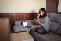 Jovem fêmea étnica com vidro de café surf internet no celular sentado no sofá na sala de casa perto do laptop — Fotografia de Stock
