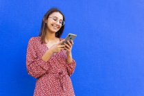 Mujer sonriente en vestido y gafas de vista de pie cerca de la pared azul y el uso de teléfono inteligente en el día - foto de stock