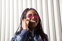 Веселая стильная молодая афроамериканка с афрокосичками в модной куртке и солнцезащитных очках говорит по мобильному телефону, стоя возле стены городского здания — стоковое фото