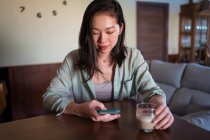 Молода етнічна жінка зі склянкою кави, що серфінгує Інтернет на мобільному телефоні за столом у домашній кімнаті — стокове фото