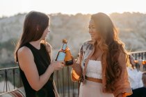 Vue latérale de jeunes amies multiethniques positives souriant brillamment et cliquetis bouteilles de bière tout en profitant de moments agréables ensemble au coucher du soleil sur la terrasse bar en Cappadoce, Turquie — Photo de stock