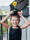 Criança alegre no avental de jardinagem com Helianthus florescente no pote na cabeça olhando para a câmera na varanda — Fotografia de Stock