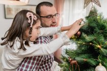Padre dando piggy indietro giro a figlia decorare abete di Natale con stella festiva mentre si prepara per la celebrazione — Foto stock