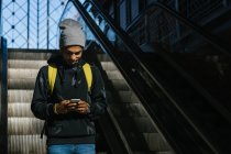 Side view of delighted African American male hipster with backpack standing on moving stairs surfing mobile phone — Stock Photo