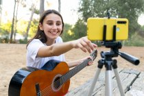 Улыбающаяся гитаристка-подросток записывает видео на современном мобильном телефоне на треноге в парке на размытом фоне — стоковое фото