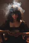 Ведьма в черном платье и с раскрашенным лицом, стоящим с чашей в комнате с паром во время духовного ритуала — стоковое фото