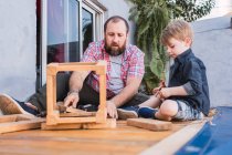 Unshaven mature dad with attentive boy measuring wooden blocks with tape while spending time on blurred background — Stock Photo