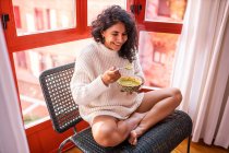 Von oben sitzt eine barfüßige lateinamerikanische Frau mit gekreuzten Beinen und geschlossenen Augen auf einem Stuhl und isst Suppe aus einer Schüssel — Stockfoto
