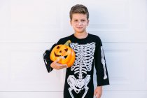 Cuerpo completo de niño preadolescente sonriente vestido de Halloween negro con esqueleto estampado de pie cerca de Jack O linterna tallada calabaza contra la pared blanca - foto de stock