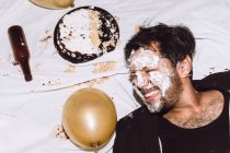 Borracho riendo macho en pastel de cumpleaños aplastado acostado cerca de botellas vacías de cerveza y globos con los ojos cerrados - foto de stock