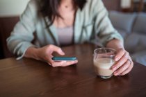 Обрезанные неузнаваемая женщина со стаканом кофе серфинг интернет на мобильный телефон за столом в доме комнате — стоковое фото