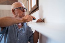 Attentive elderly male in eyeglasses with manual screwdriver screwing shelf to wall in house room — Stock Photo