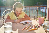 Позитивная девушка с темными волосами в повседневной одежде сидит за столом с маркерами и рисует на бумаге на террасе в солнечный день — стоковое фото