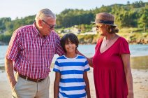 Full body of positive senior couple embracing boy while standing looking at camera on sandy coast of river in countryside — Stock Photo