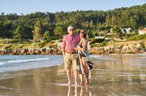 Улыбающаяся босиком пожилая пара в солнечных очках, стоящая на мокром песчаном пляже и наслаждающаяся солнечным днем — стоковое фото