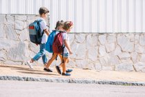 Вид сбоку школьника с рюкзаком, который разговаривает с подругами, прогуливаясь по плиточной тротуарной плитке на фоне каменной стены при солнечном свете — стоковое фото