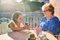 Счастливая внучка сидит за столом и показывает фотографии на смартфоне улыбающейся бабушке, сидя на террасе в солнечный день — стоковое фото