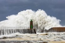 D'énormes vagues de mer mousseuses s'écrasent contre un brise-lames pierreux avec une vieille tour de phare contre un ciel nuageux bleu à Port of Viavelez dans les Asturies Espagne — Photo de stock