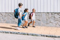 Side view of schoolboy with backpack speaking with female friends while strolling on tiled pavement against stone wall in sunlight — Stock Photo