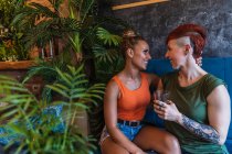 Contenuto giovane donna tatuata con mohawk e bevanda abbracciando fidanzata lesbica mentre si guardano sul divano in casa — Foto stock