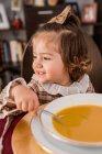 Очаровательный ребенок с луком на каштановых волосах и ложкой, смотрящий в сторону тарелки супа из пюре для сквоша в доме — стоковое фото