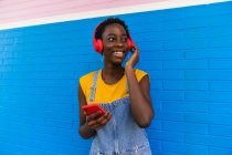 Encantada mujer afroamericana disfrutando de canciones en auriculares de la lista de reproducción en el teléfono inteligente contra la pared colorida - foto de stock