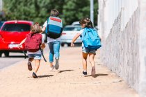 Back view of anonymous schoolkids with backpacks running on tiled walkway in sunny town on blurred background — Stock Photo