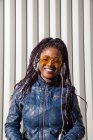 Feliz joven afroamericana femenina con trenzas afro vestida con chaqueta azul y gafas de sol elegantes disfrutando de la música a través de auriculares mientras se enfría a la luz del sol contra la pared rayada - foto de stock