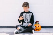 Full body of cheerful boy in black skeleton costume with painted face and carved Halloween pumpkin using on mobile phone while sitting near white wall on street — Stock Photo