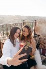Hochwinkel ethnisch glücklicher Freundinnen, die auf Kissen sitzen, während sie Selbstporträts auf dem Handy am Tisch mit Sofortfotokamera und Cocktail auf der Terrasse in Kappadokien, Türkei, machen — Stockfoto