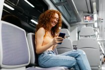 Цікава жінка з кучерявим волоссям в рваних джинсах текстових повідомлень на мобільний телефон під час поїздки в поїзді вдень — стокове фото