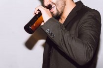 Crop allegro maschio irriconoscibile con gli occhi chiusi bere birra dalla bottiglia durante la festa contro sfondo bianco — Foto stock