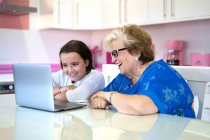 Nieta sonriente y abuela sentadas en la mesa y viendo videos en la computadora portátil en la cocina ligera en el apartamento - foto de stock