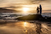 Повернення до нерозпізнаної молодої жінки, що стоїть на березі з дошкою для серфінгу перед тим, як сідати в море під час заходу сонця на пляжі в Астурії (Іспанія). — стокове фото