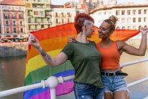 Mulher tatuada legal com mohawk e bandeira LGBTQ abraçando namorada com olhos fechados contra o canal na cidade — Fotografia de Stock