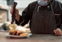Неузнаваемый мужчина-повар в стерильной маске, украшающий вкусные венские вафли шоколадным соусом из бутылки на кухне ресторана — стоковое фото