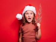 Cute astonished little girl in casual clothes and Santa hat holding fir tree twig and looking at camera against red background — Stock Photo
