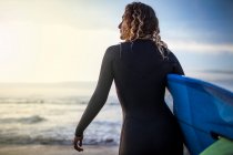 Вид сзади на неузнаваемую молодую женщину, стоящую на берегу с доской для серфинга, прежде чем попасть в море во время заката на пляже в Астурии, Испания — стоковое фото