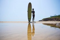 Vue arrière de l'athlète afro-américaine avec planche de surf admirant l'océan depuis le rivage sablonneux sous un ciel bleu nuageux — Photo de stock