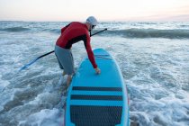 Visão traseira do surfista masculino irreconhecível em roupa de mergulho e chapéu carregando prancha de remo e entrando na água para surfar na costa — Fotografia de Stock