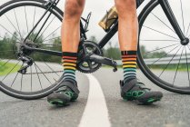 Nivel del suelo del atleta masculino sin rostro en zapatillas de ciclismo y calcetines a rayas de pie en la carretera con bicicleta - foto de stock